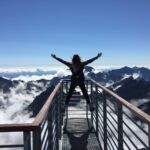person standing on hand rails with arms wide open facing the mountains and clouds